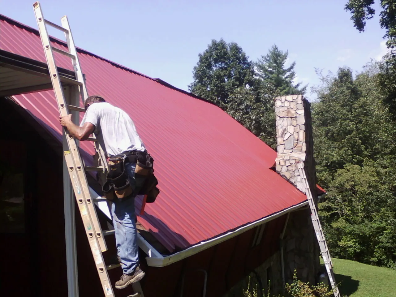 Roofer on a ladder beside a red metal roof and stone chimney — Soffit & Fascia Repair by Roofing Brothers in Powers Lake